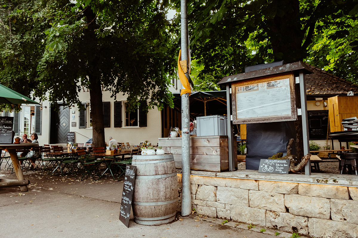 Eingang zu einem Biergarten eines Wirtshauses, in welchem Leute an den Tischen sitzen.