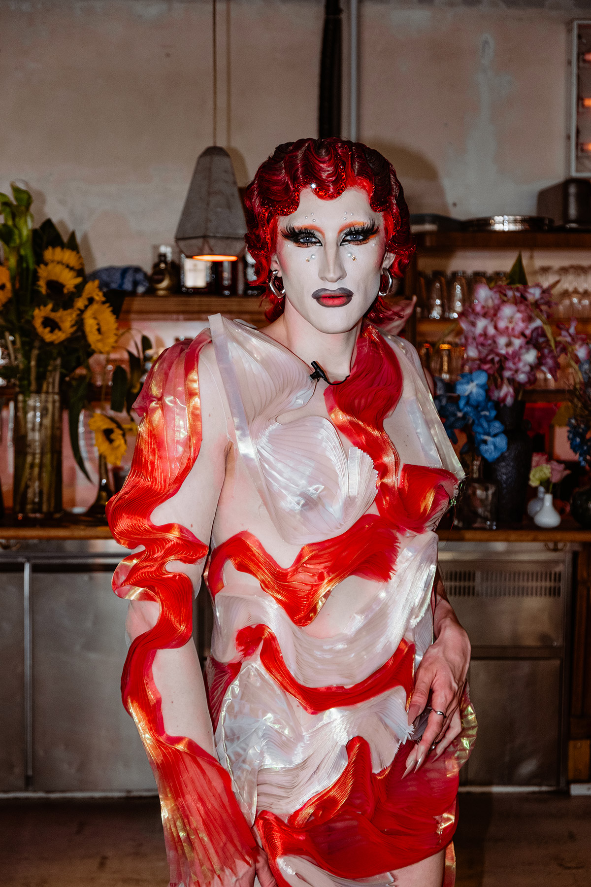 Drag-Queen steht in einem Raum, mit vielen Blumen im Hintergrund.