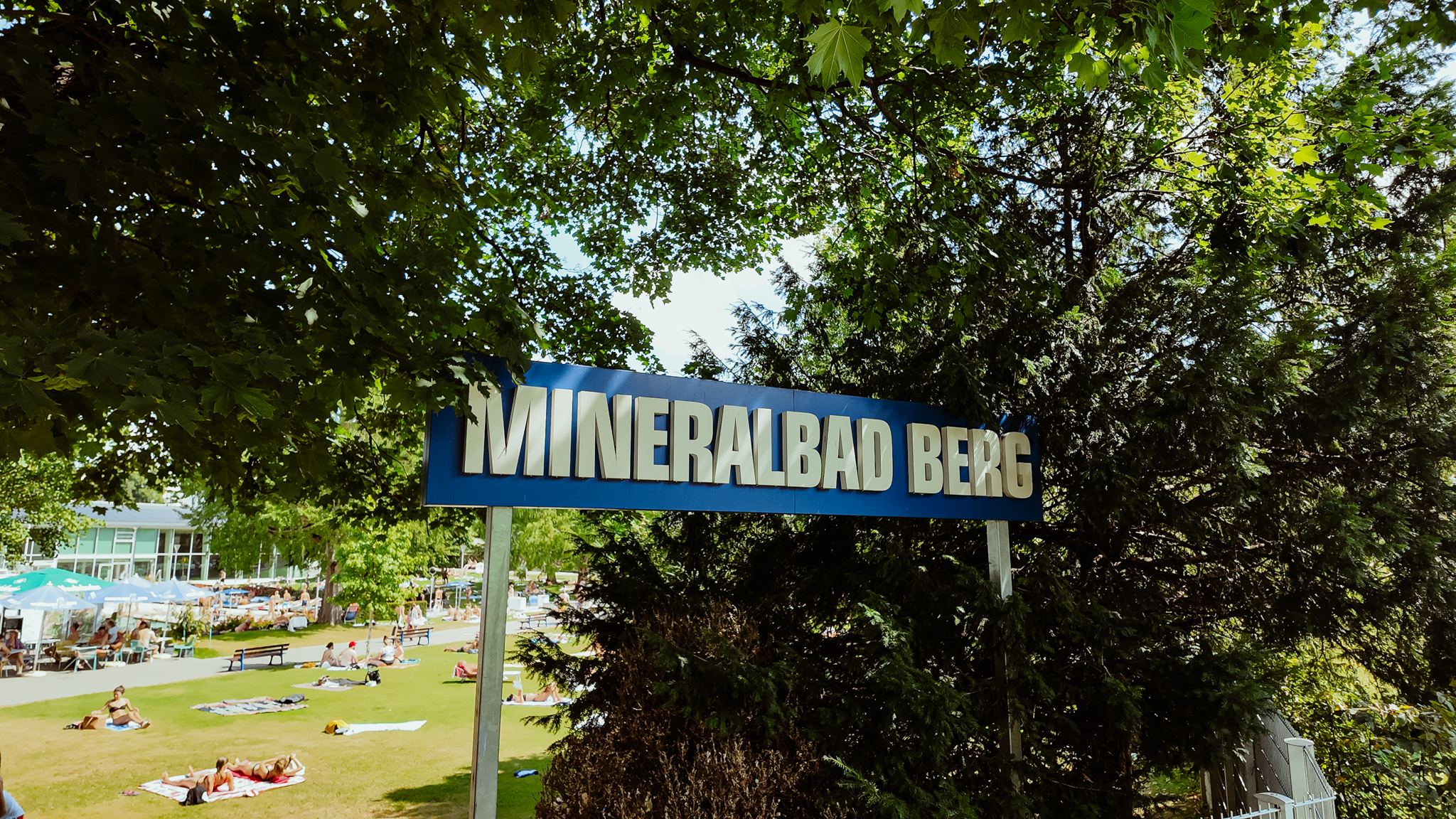 Schild mit der Aufschrift "Mineralbad Berg" zwischen Büschen/Bäumen. Im Hintergrund sonnen sich Menschen auf einer Wiese.