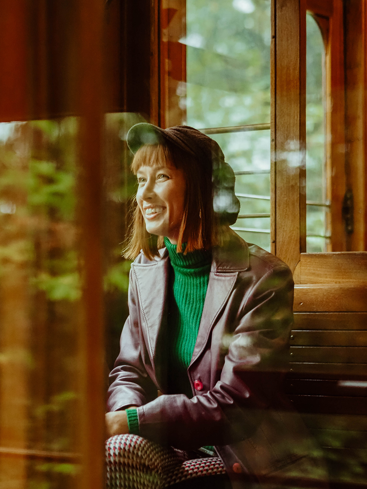 Alisa Scetinina alias Gaisma sitzt lachend in der Seilbahn, das ist durch das Fenster aufgenommen, in dem sich der Wald spiegelt. Sie trägt einen grünen Rollkragenpullover und darüber ein violettes Jacket, auf ihrem Kopf sitzt eine Schiebermütze.