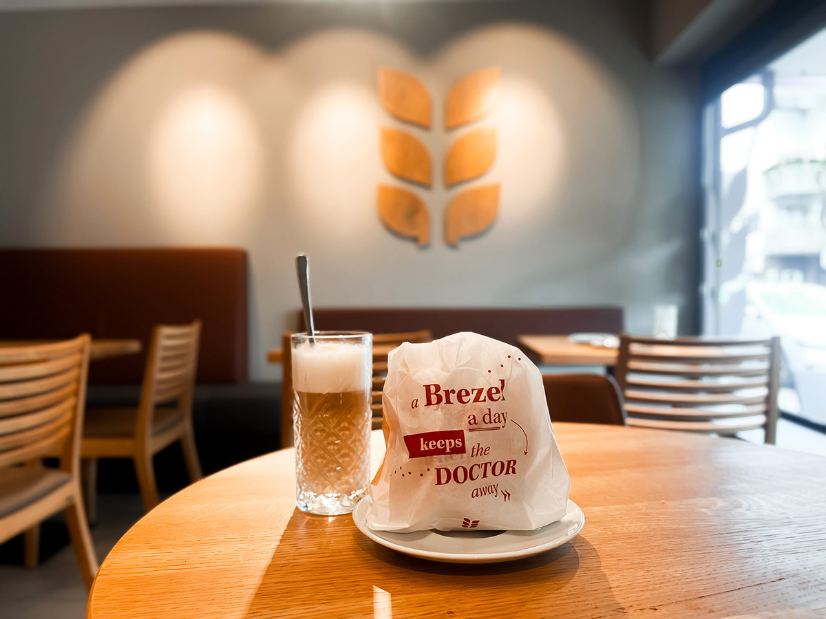 Aufnahme im Bäcker Schultheiß in Leinfelden unweit der Haltestelle Neuer Markt: Auf einem Tisch steht eine Tüte mit einer Brezel und der Aufschrift "a Brezel a day keeps the doctor away", daneben steht ein Kaffee Latte mit viel Milchschaum in einem durchsichtigen Longdrink-Glas