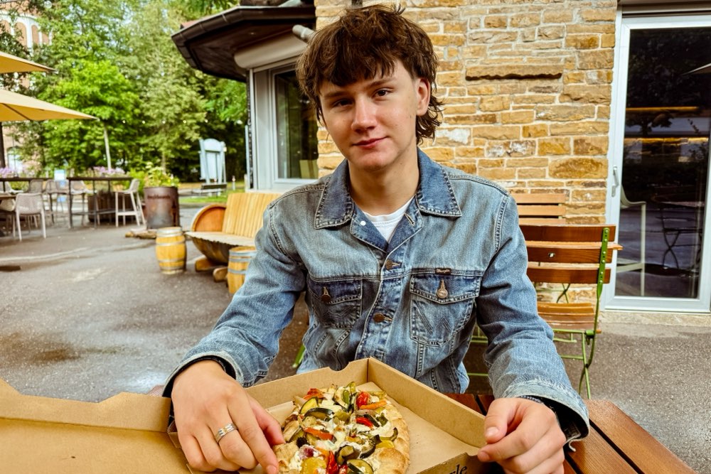 Junger Mann mit Jeansjacke sitzt an einem Tisch und isst eine Dinnete 